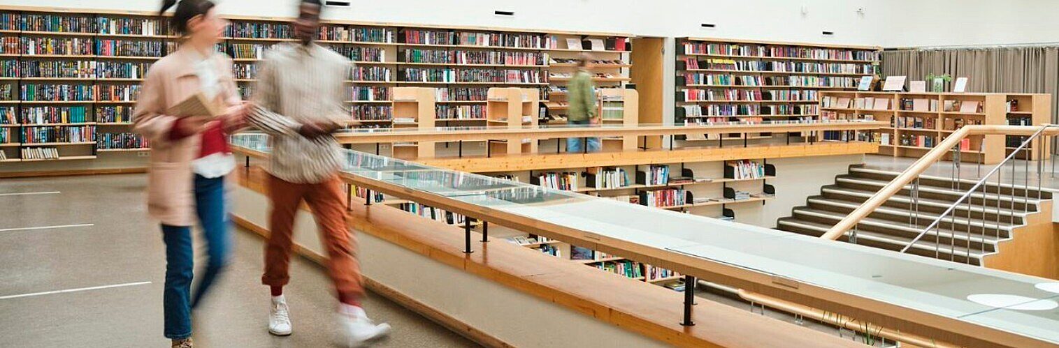 Two people walking with motion blur through a modern, multi-level library lined with bookshelves full of books.