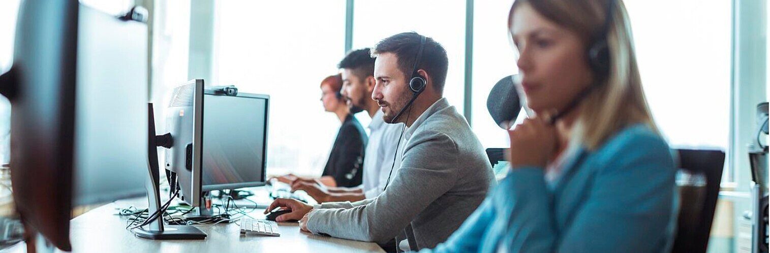 Row of customer service agents wearing headsets and working on computers in a busy call centre office.