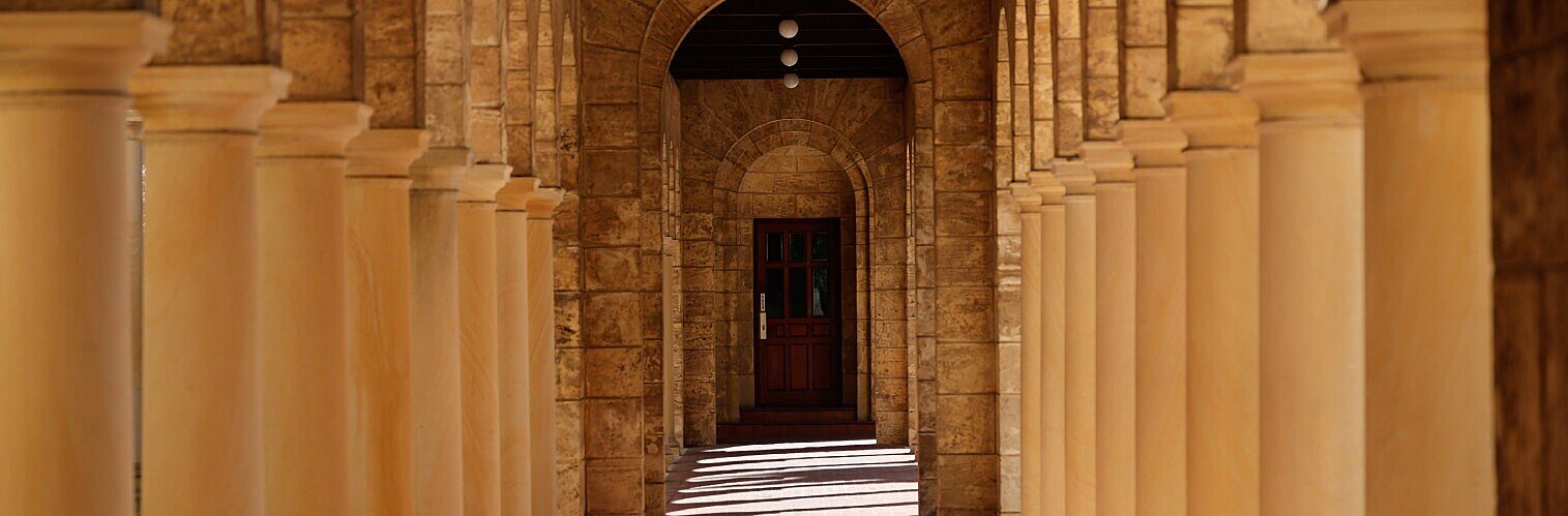 A sunlit stone colonnade with rows of columns casting long shadows, leading towards an arched doorway at the far end.