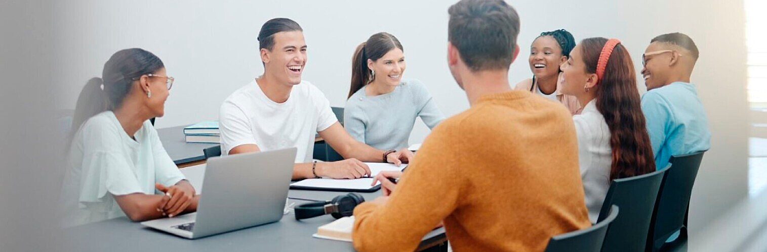 Group of diverse young people sitting around a table, actively engaged in a cheerful discussion or collaborative session in a bright room.