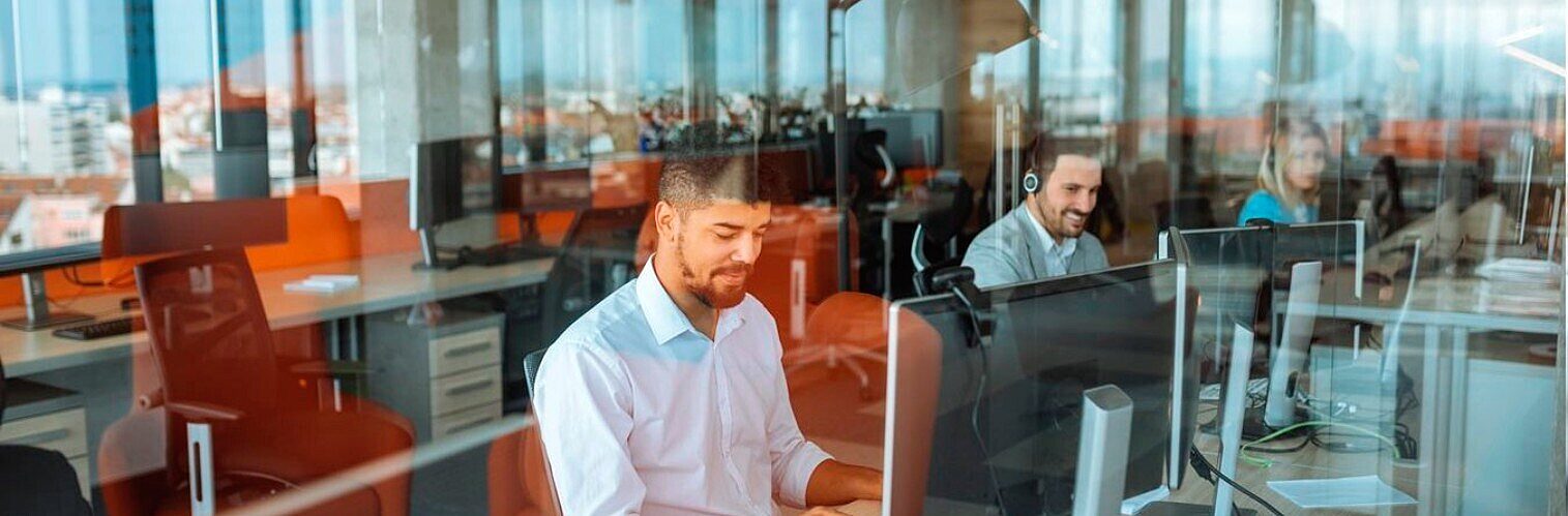 Man working at a computer in a busy, modern open-plan office viewed through a glass partition.