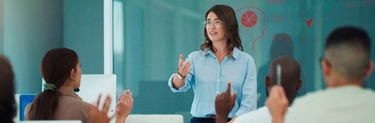 Female lecturer or tutor teaching a class, gesturing towards a chalkboard with diagrams while students listen.