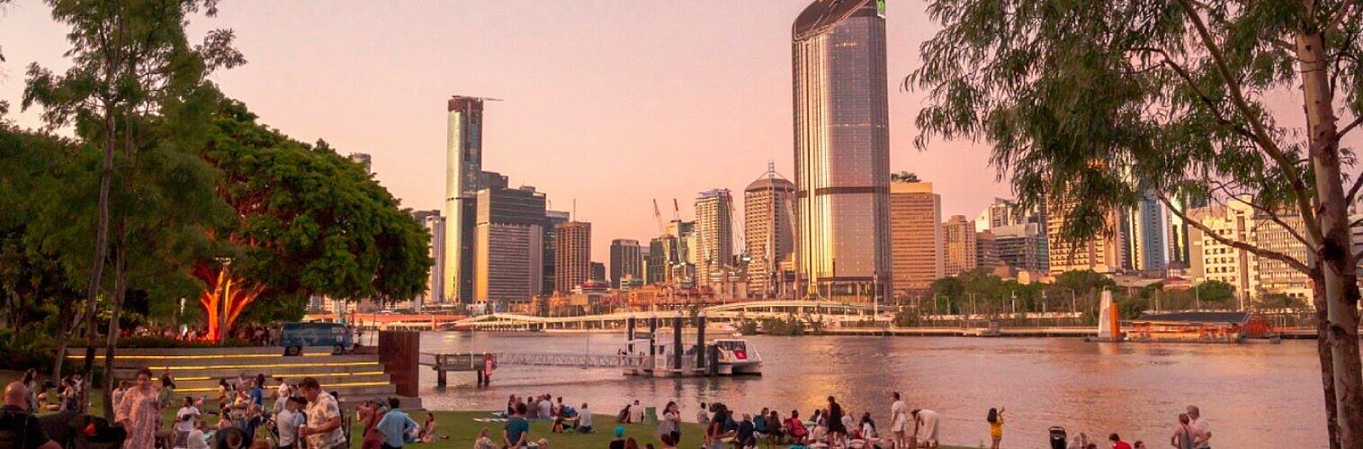 Brisbane city skyline viewed across the river from a busy park at sunset, with people relaxing on the grass.
