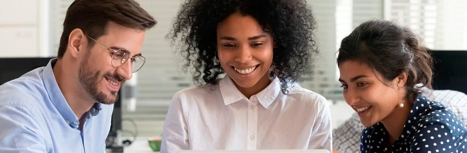 Three diverse colleagues smiling and collaborating closely while looking at a laptop screen in an office.