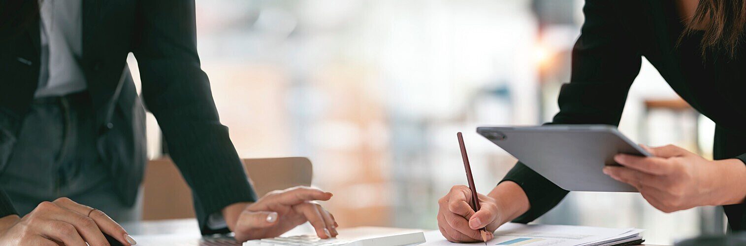 Two colleagues analysing information together at an office desk, using a tablet, paper documents, and a pencil, representing strategic work on university financial challenges.