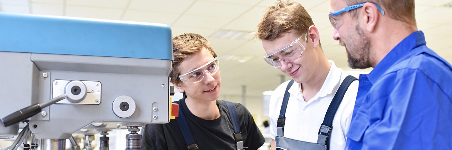 Instructor teaching two young male apprentices or students using machinery in a workshop, all wearing safety glasses.