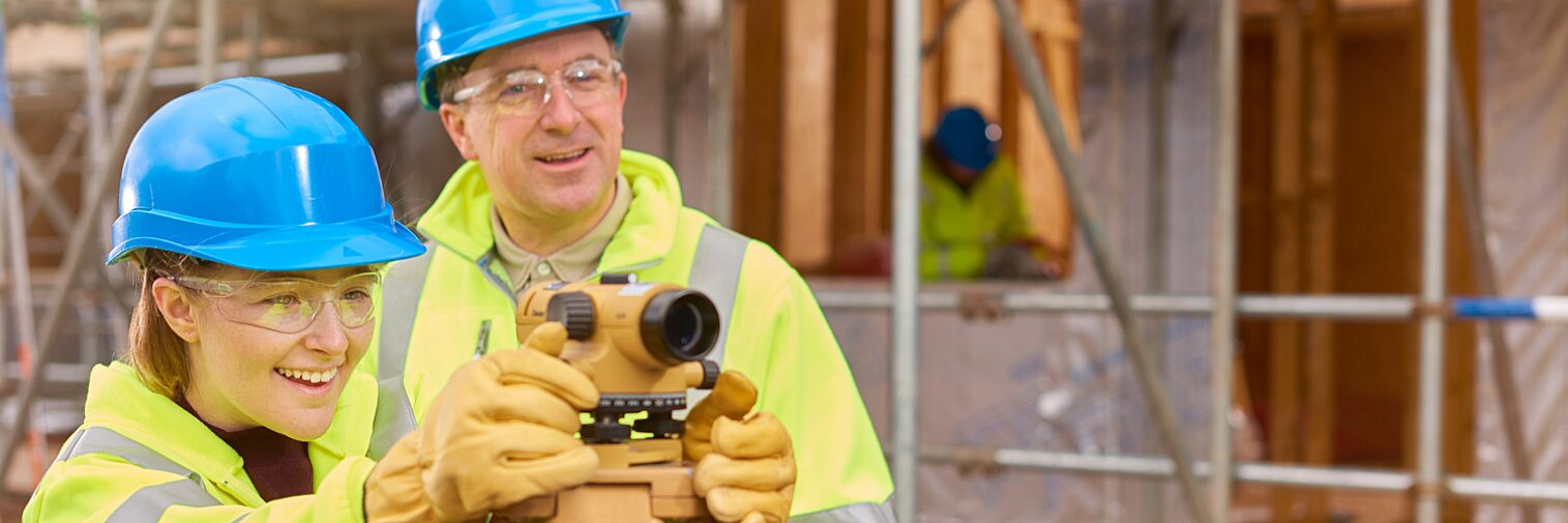 Apprentice surveyor learning to use a level tool on a construction site under the supervision of a male instructor.