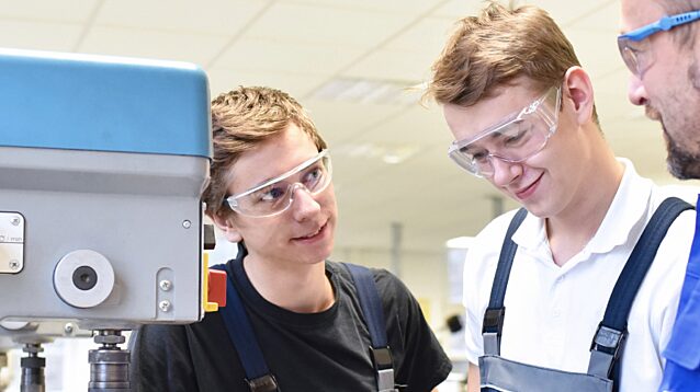 Instructor teaching two young male apprentices or students using machinery in a workshop, all wearing safety glasses.