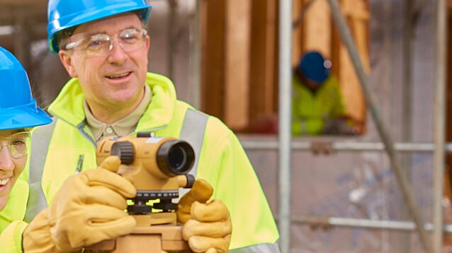 Apprentice surveyor learning to use a level tool on a construction site under the supervision of a male instructor.