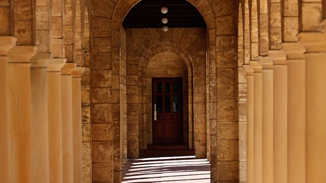 A sunlit stone colonnade with rows of columns casting long shadows, leading towards an arched doorway at the far end.
