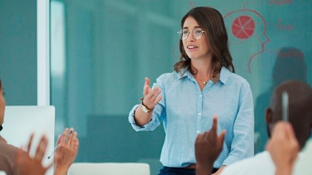 Female lecturer or tutor teaching a class, gesturing towards a chalkboard with diagrams while students listen.