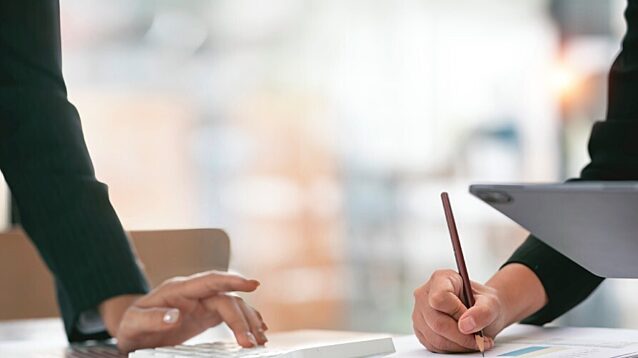 Two colleagues analysing information together at an office desk, using a tablet, paper documents, and a pencil, representing strategic work on university financial challenges.