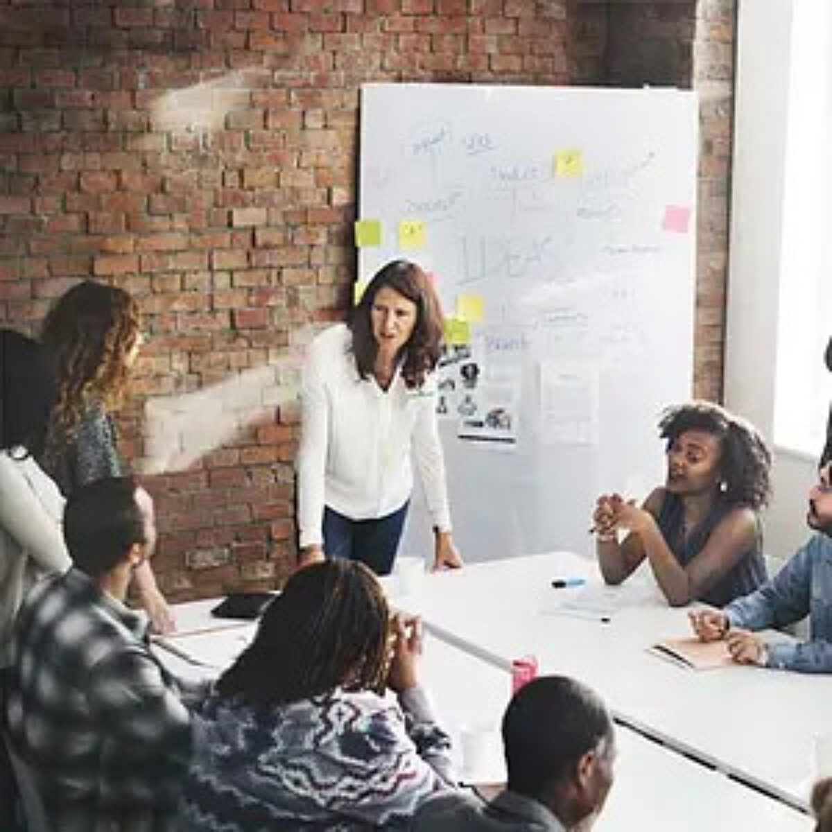 A diverse team of colleagues collaborating around a meeting table in a modern office with a brick wall. One woman stands presenting next to a whiteboard covered in ideas and sticky notes, while others listen and engage.