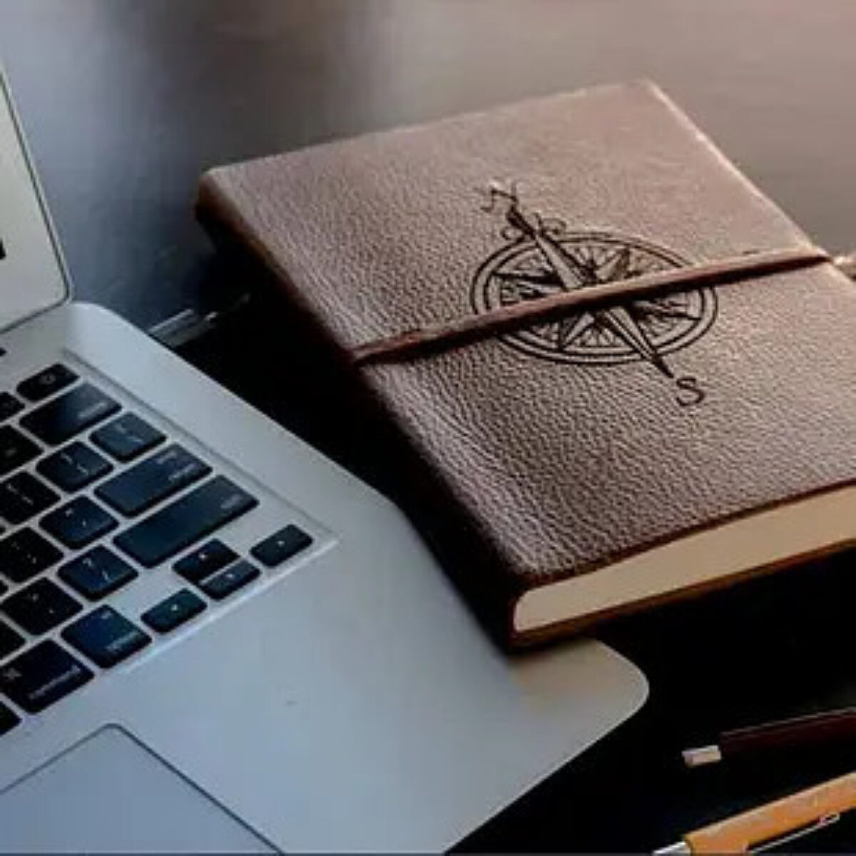 Desk workspace featuring a silver laptop positioned next to a closed, brown leather-bound journal embossed with a compass rose design and secured with an elastic strap. Pens are visible nearby on the dark surface.