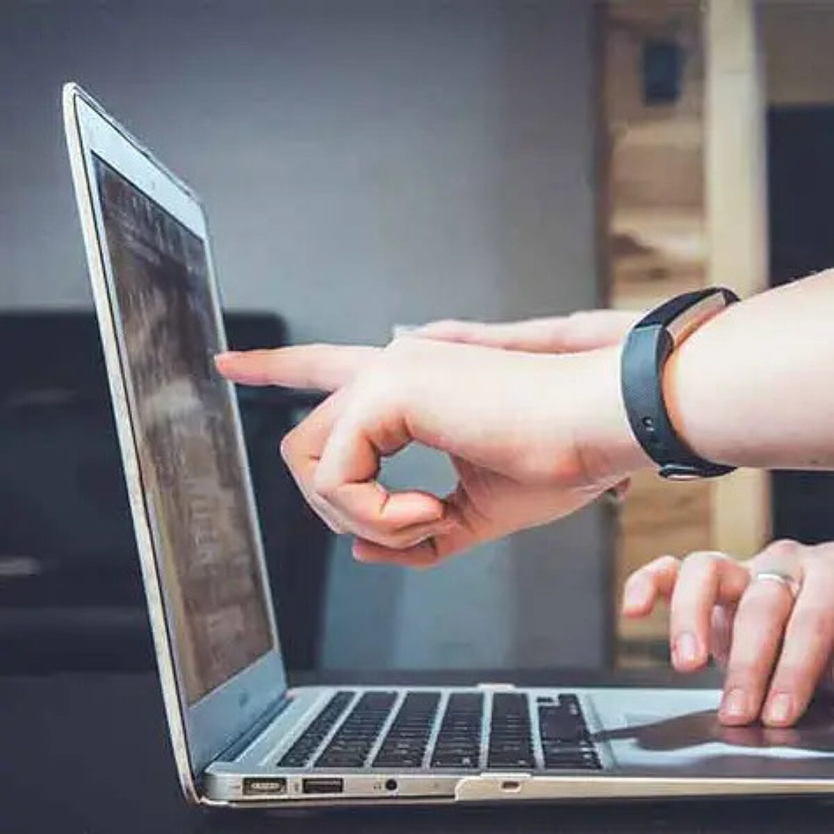 Close-up of two people's hands interacting with a silver laptop computer. One hand points firmly at the screen, while the other rests near the keyboard and trackpad, suggesting teamwork, discussion, or collaboration while reviewing content.