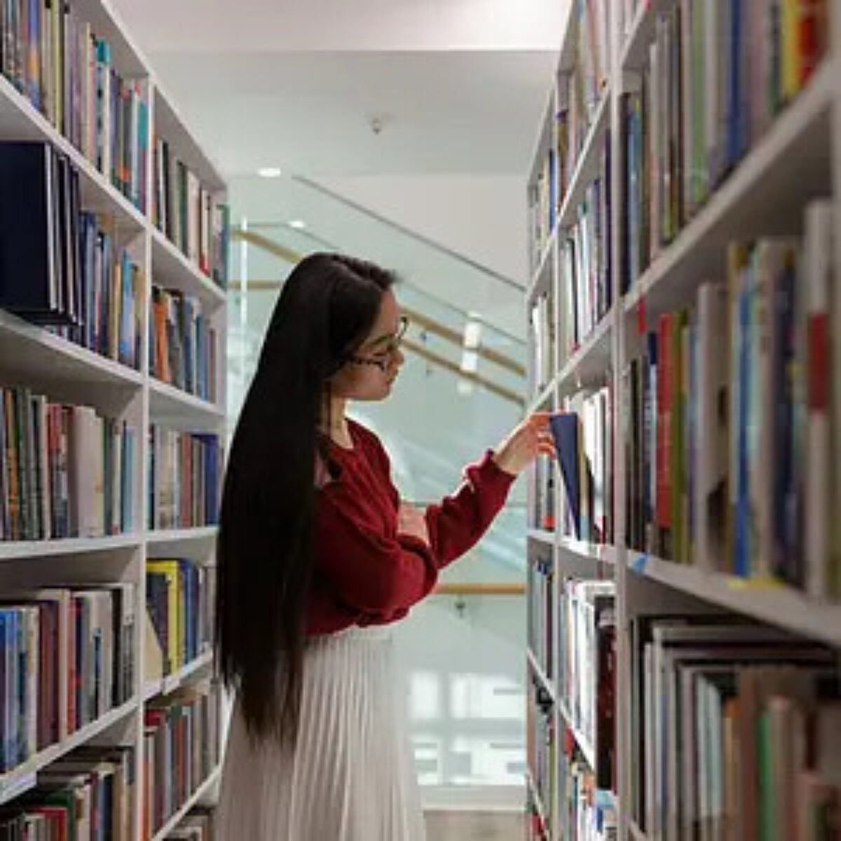 Young woman with long dark hair and glasses, wearing a red jumper and white skirt, standing between tall library bookshelves and carefully selecting a blue book from the shelf.