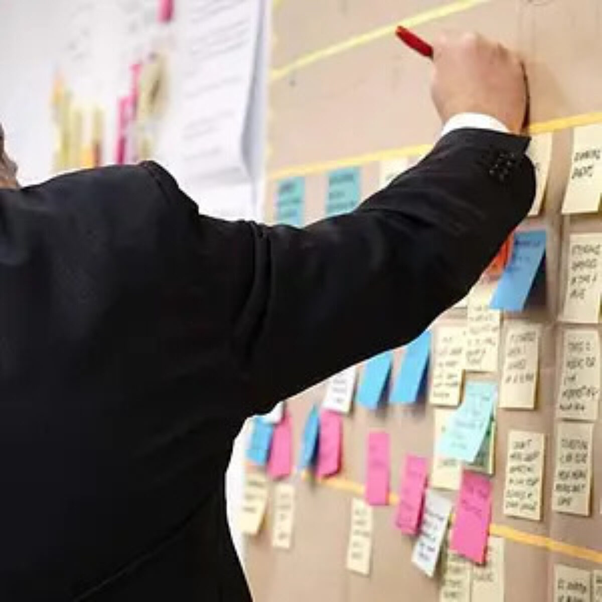 Business person in a suit jacket, seen from behind, writing with a red pen on a large board covered with colourful sticky notes arranged in columns, suggesting a planning, brainstorming, or project management session.
