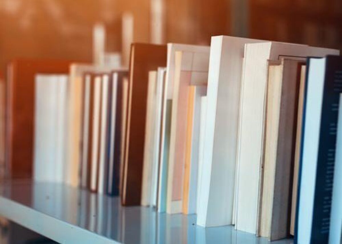 Close-up side view of a row of books with varied spines standing vertically on a shelf. Warm lighting illuminates the background, evoking a library, study, or reading atmosphere.