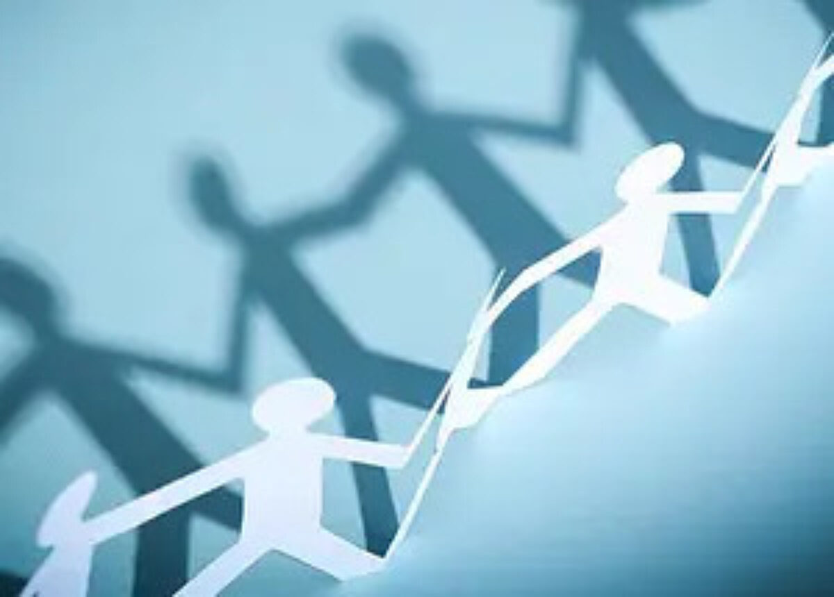 Chain of white paper cut-out people holding hands, stretching diagonally across a blue surface and casting strong shadows. Represents concepts like teamwork, unity, connection, or community.